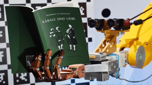 Ein Roboter hält ein Buch in der künstlichen Hand und liest mit seinen zwei Kameraaugen.